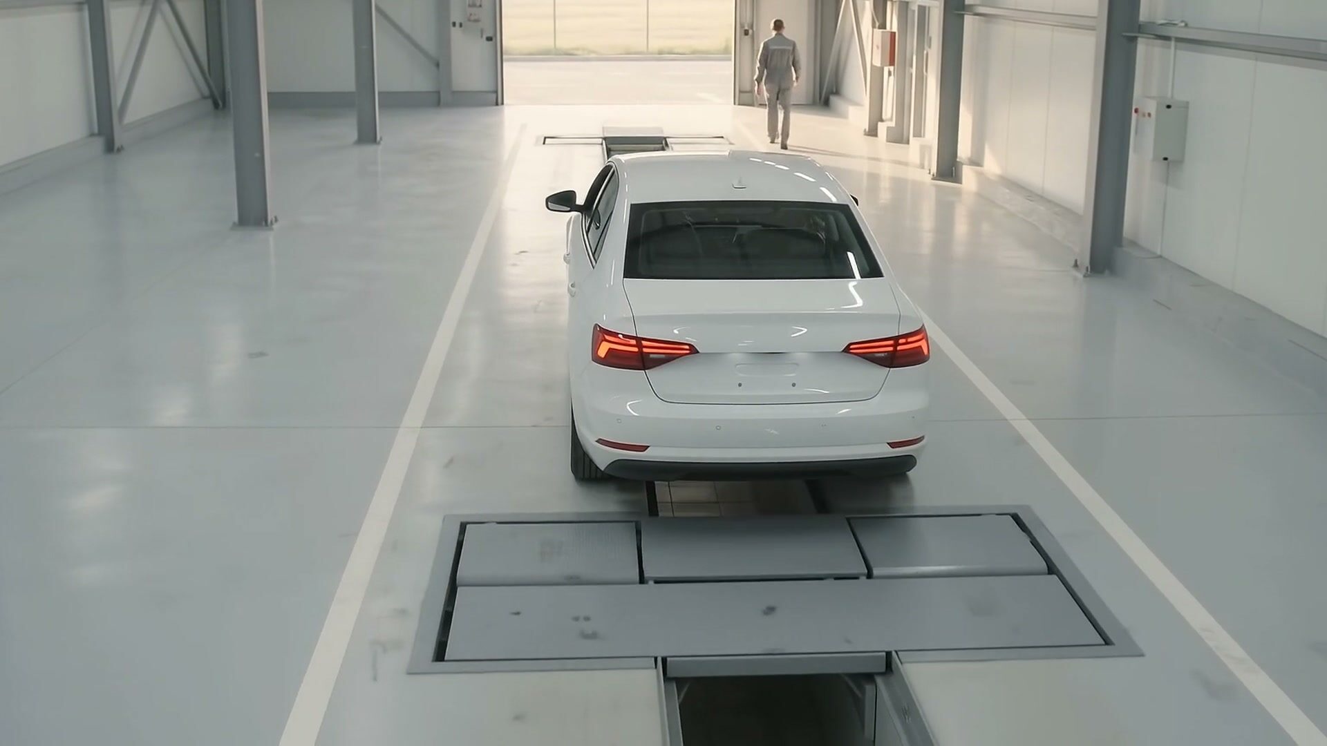 A car driving along its inspection lane over the pit, seen from behind, with an inspector far off by the open door